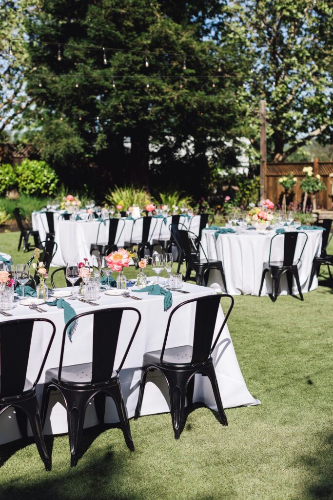 Outdoor dining setup with round tables draped in white cloths, black chairs, and floral centerpieces on a grassy lawn, perfect for a Napa Valley wedding.