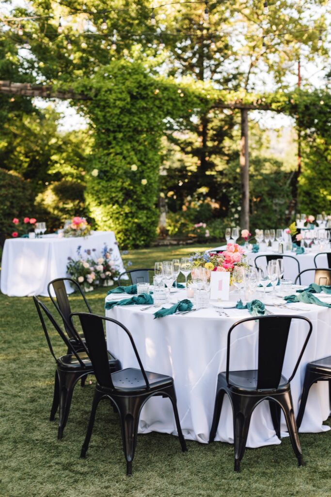 Experience a Napa Valley wedding in an enchanting outdoor garden setting, where round tables draped in white cloth showcase vibrant floral centerpieces and green napkins. Surrounded by black chairs and lush greenery, this picturesque scene captures the essence of a wine country celebration.