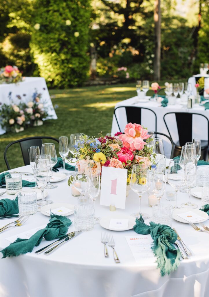Napa Valley wedding table setting features a white tablecloth, floral centerpiece, glassware, cutlery, and green napkins. A numbered card rests amid the elegance. Lush greenery from the valley is visible in the background, enhancing the serene ambiance.