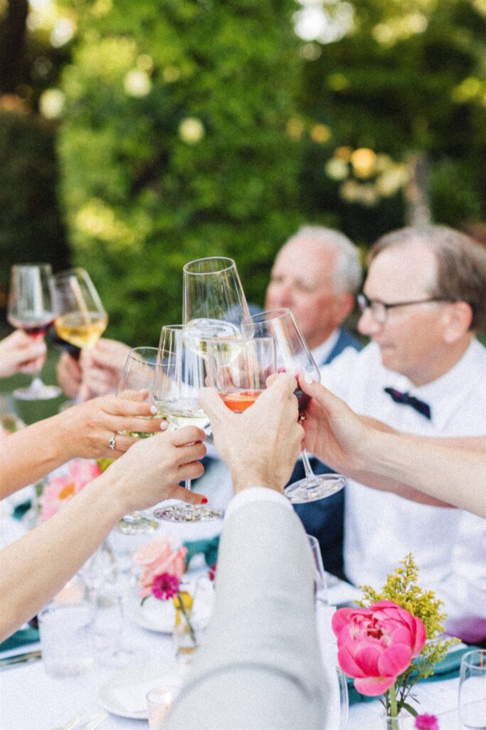 A group of people clinking wine glasses at an outdoor table, surrounded by flowers and greenery, celebrates a picturesque Napa Valley wedding.