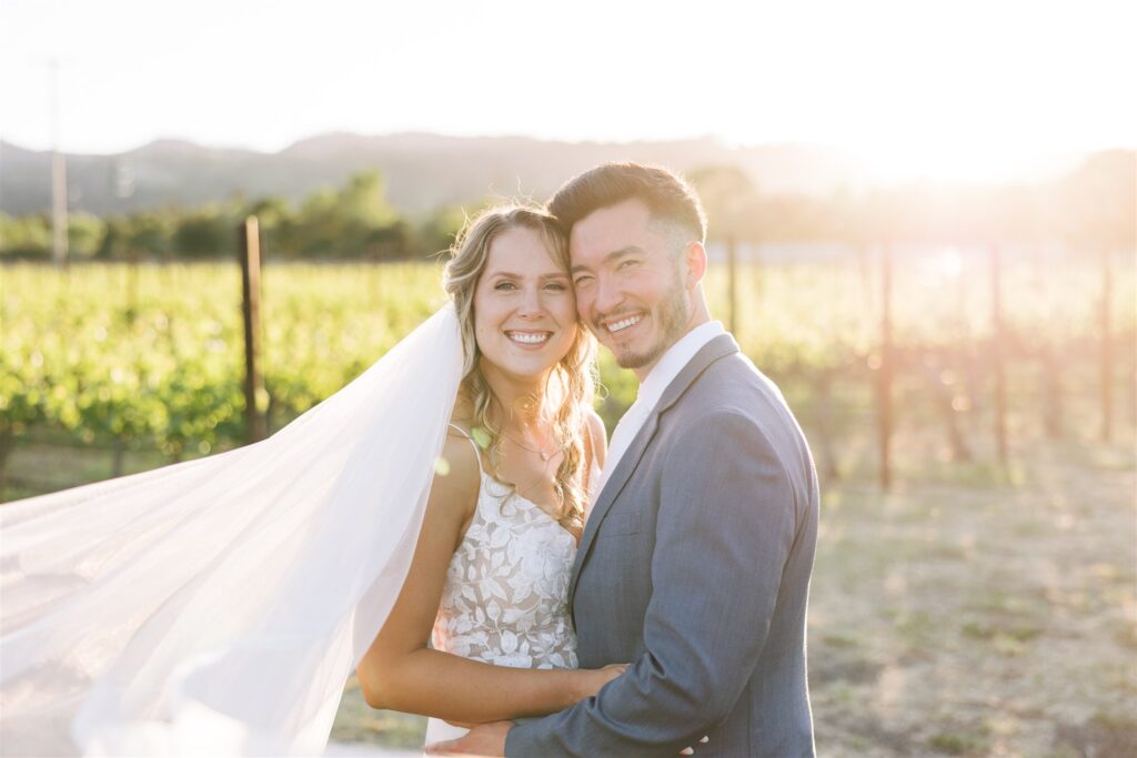 A bride and groom smile while standing in a sunlit Napa Valley vineyard, with the bride's veil flowing in the breeze.