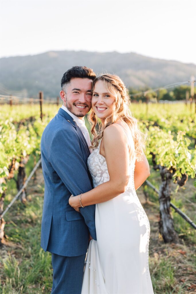 A couple in formal attire embraces in a picturesque Napa Valley vineyard, with rolling hills in the background under a clear sky, capturing the essence of a perfect wedding day.
