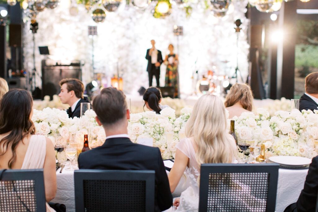 An elegantly dressed crowd seated at tables decorated with white flowers at a wedding reception, listening to speakers on a stage.