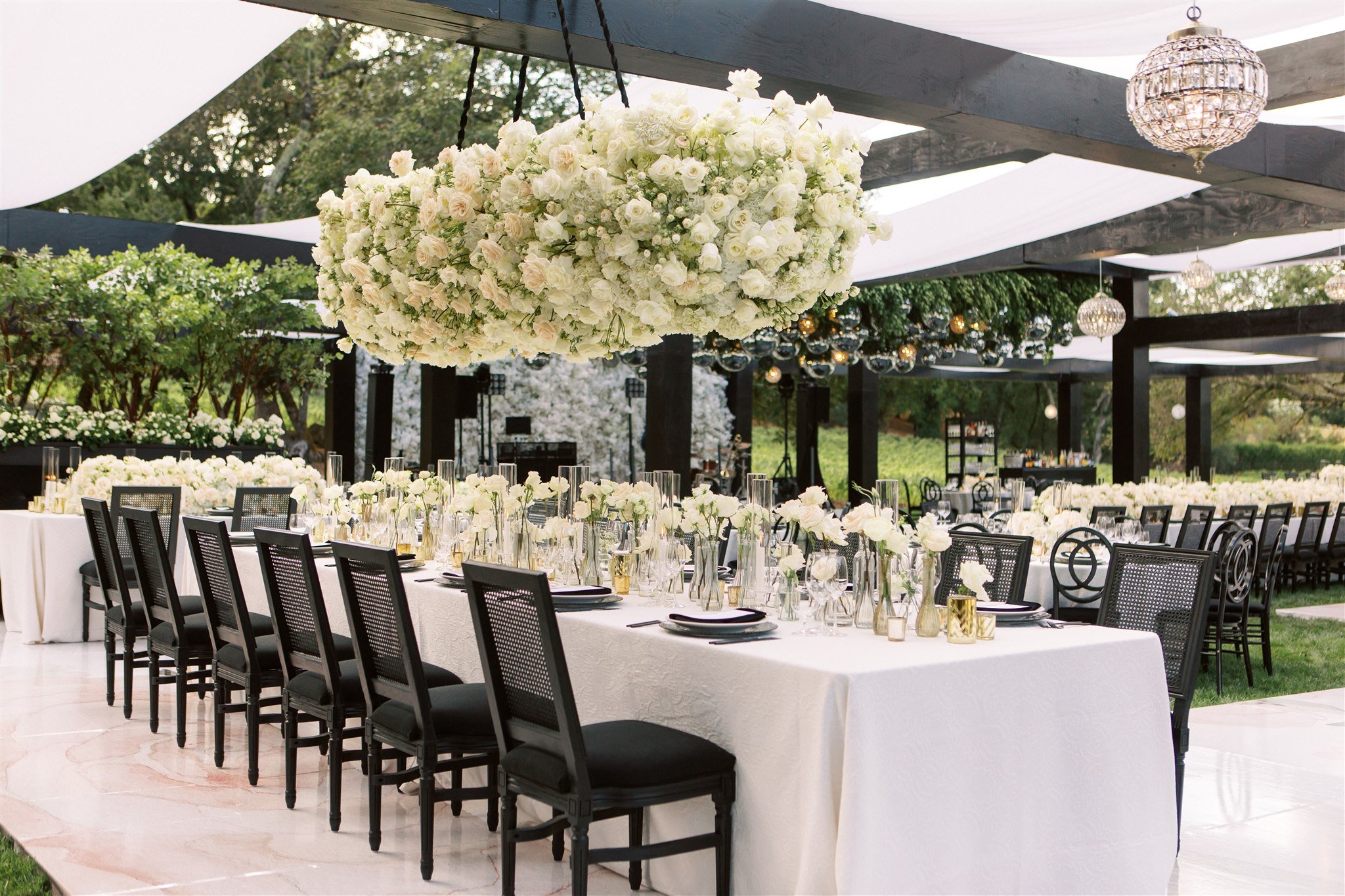 Elegant outdoor wedding reception setup with long tables, white floral centerpieces, black chairs, and large flower arrangement overhead. Tables are covered with white linens.