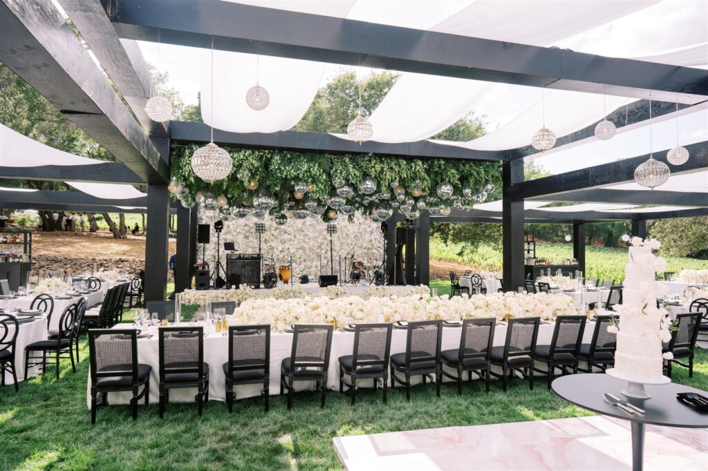 Outdoor reception setup with long dining tables, white tablecloths, and floral arrangements under a black pergola adorned with greenery and hanging orbs. A tiered white cake is in the foreground.
