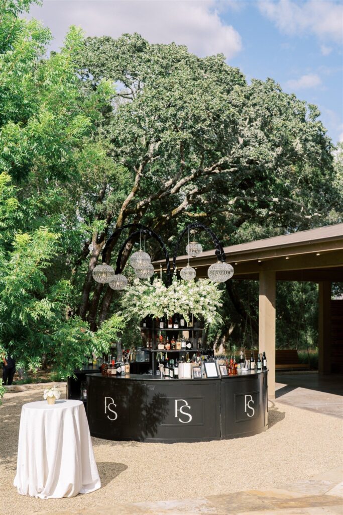 A black outdoor bar stands on a gravel patio under a large tree, with tables nearby. Glasses and drinks are arranged on the bar.