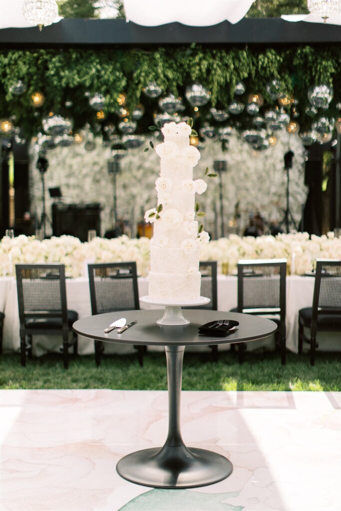 A tall, white, floral-decorated wedding cake on a round black table. Background features a flower-filled banquet setting with hanging greenery and lights.