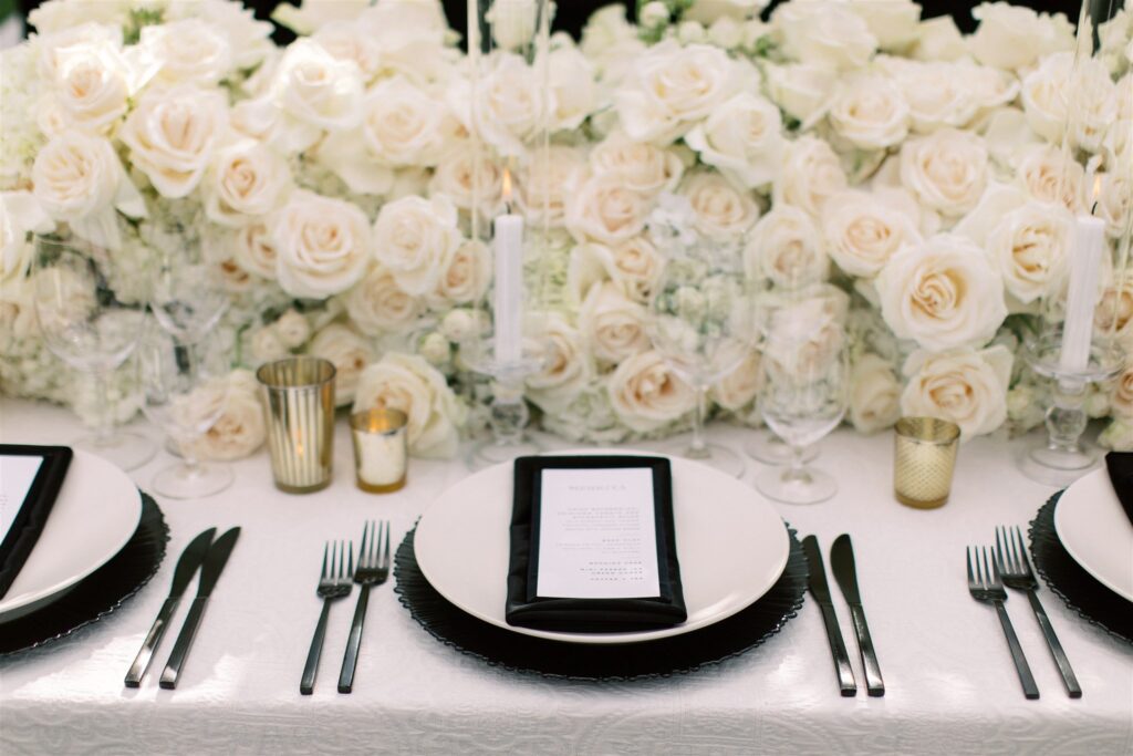 Elegant table setting with white plates, black napkins, and gold accents. Lush white and pale pink roses as a centerpiece.