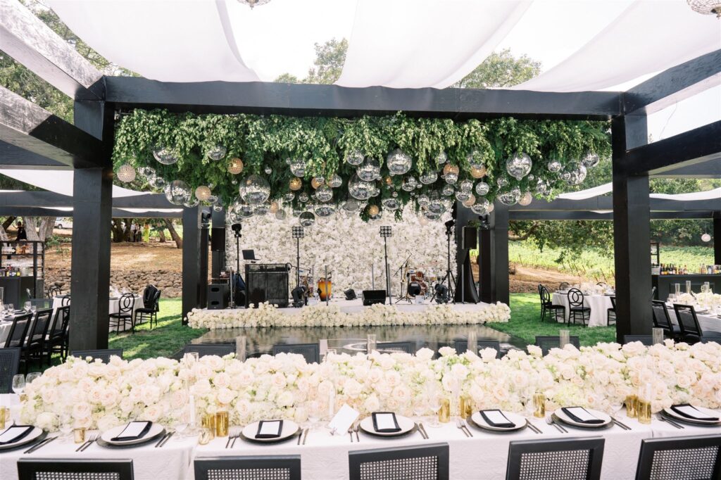 Elegant outdoor wedding setup with a floral-covered stage, hanging glass ornaments, and a long table decorated with white flowers and place settings.