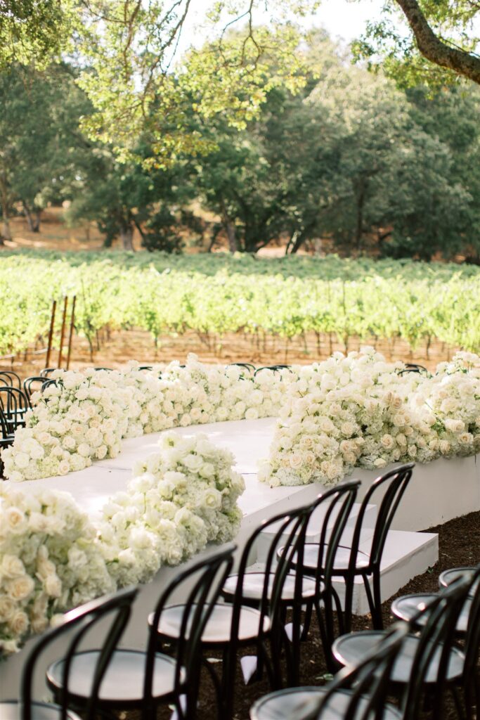 Outdoor wedding setup with chairs arranged around a floral aisle decorated with white roses, set in a vineyard with green trees and grapevines in the background.