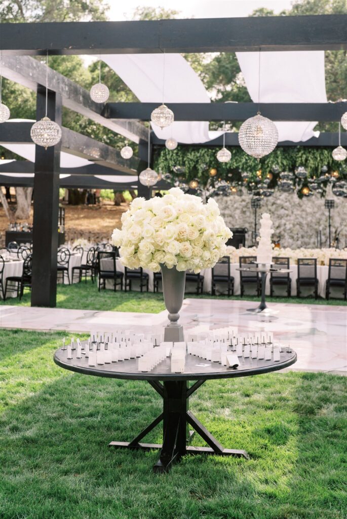 Decorative table with a white rose centerpiece, surrounded by place cards, set on grass under hanging lights and fabric drapes.