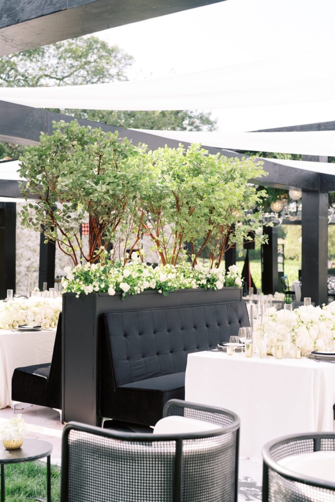 Elegant outdoor event setup with black booths, white floral arrangements, and greenery. Black chairs surround white-clothed tables under a pergola.