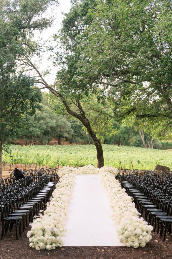 Outdoor wedding aisle lined with white flowers, surrounded by black chairs. Green trees and vineyard visible in the background.