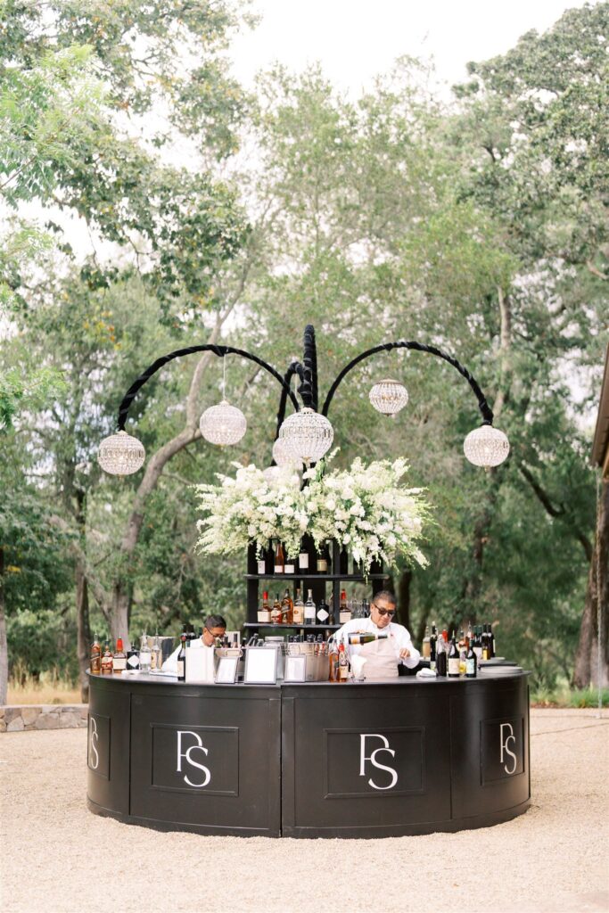 Outdoor bar setup with two bartenders, black counter with "FS" logo, adorned with white flowers and hanging crystal lights, surrounded by trees.