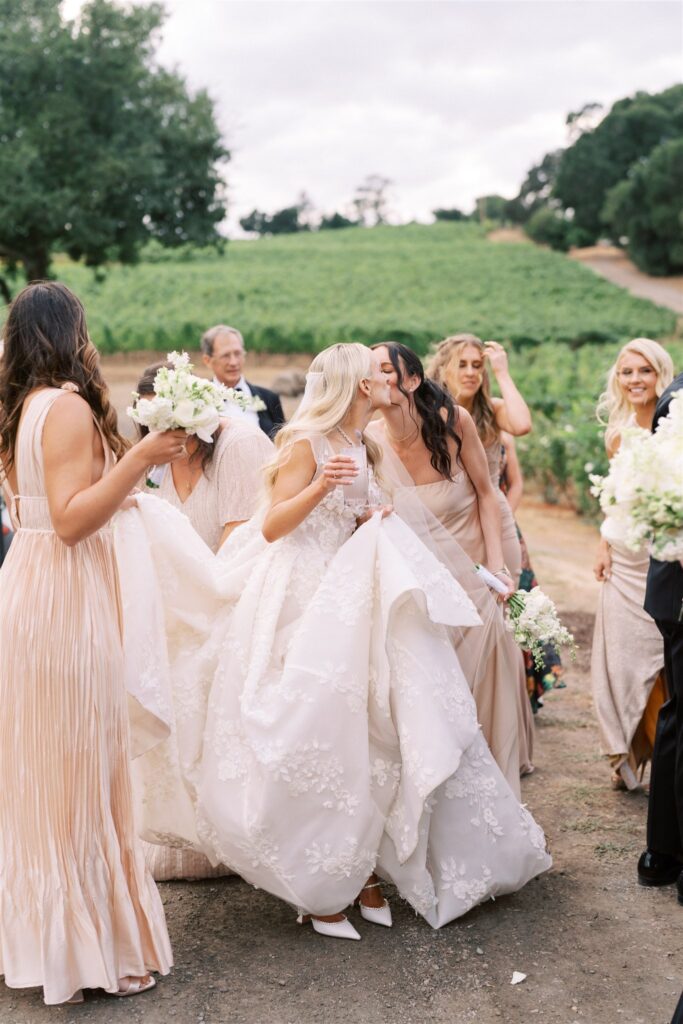A bride in a white gown shares a kiss with a woman in beige amidst a group of people in formal attire outdoors.