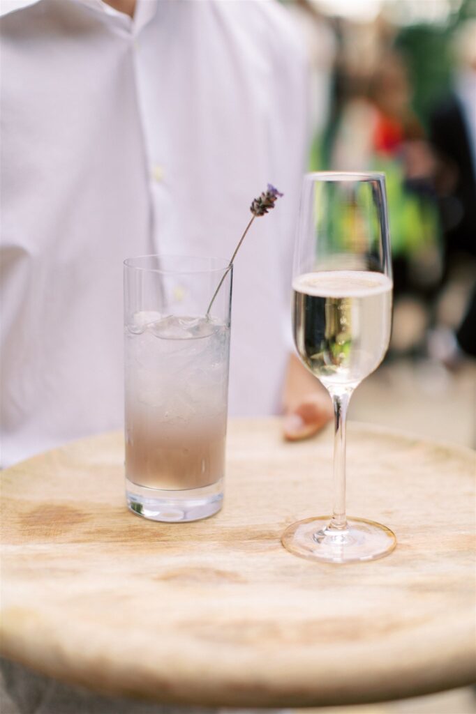 A person holding a wooden tray with a tall glass of iced lavender drink and a champagne flute.