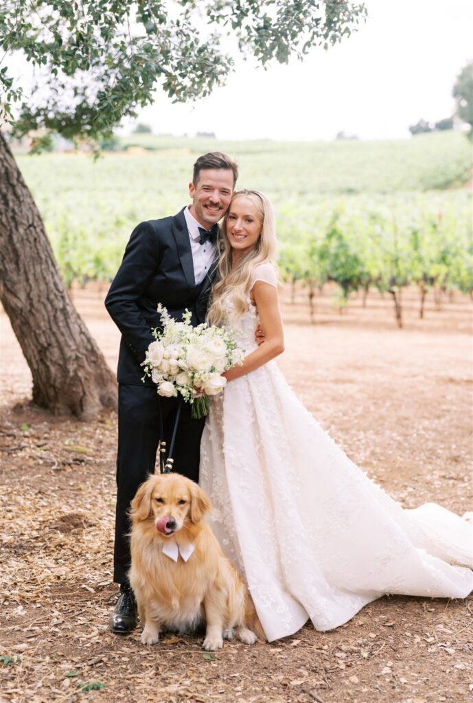 Bride and groom stand smiling with their dog in a vineyard. The bride holds a bouquet, and the dog wears a bow tie.