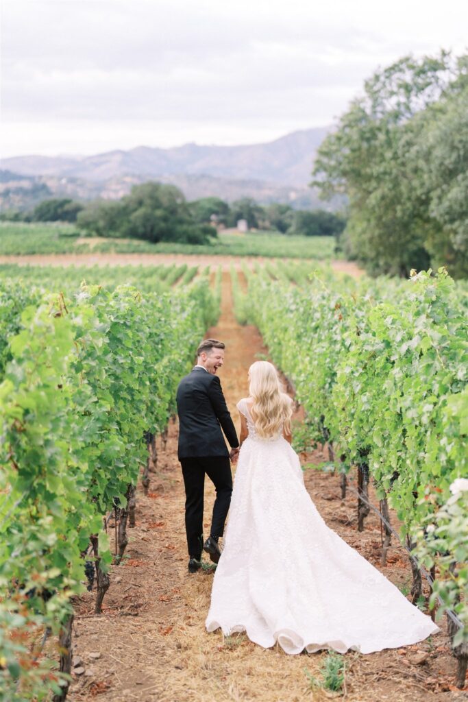 A couple in wedding attire walks hand-in-hand down a vineyard path, surrounded by rows of grapevines with mountains in the background.