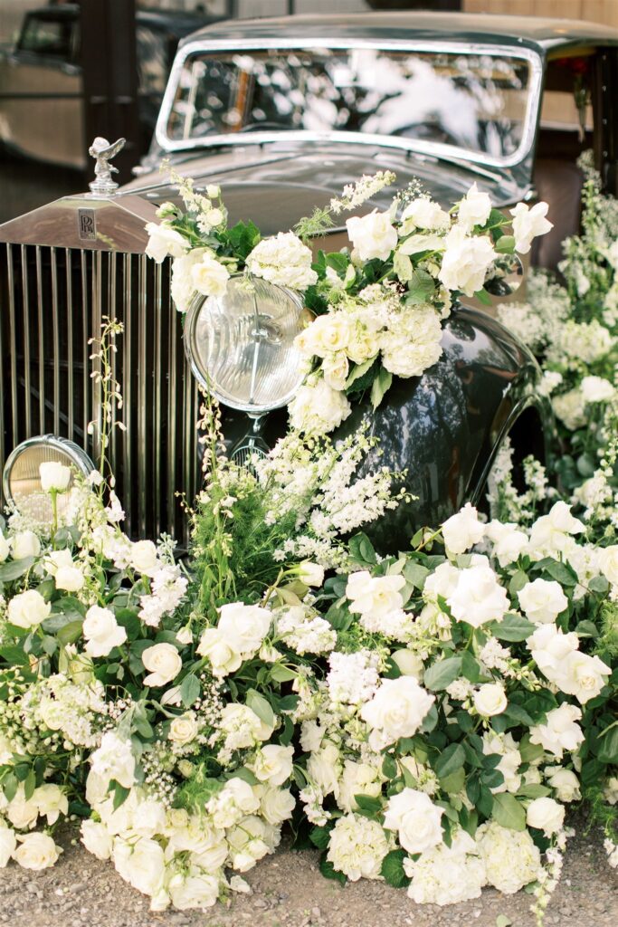 A vintage car adorned with an abundance of white roses and greenery, surrounded by floral arrangements.