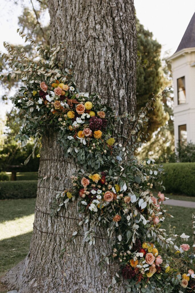 Floral arrangement with roses and greenery wrapped around a tree trunk, outdoors near a building.