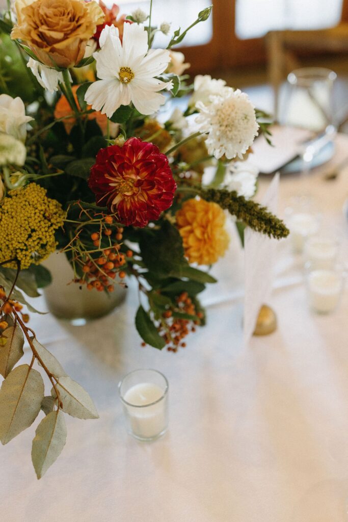 Floral arrangement with red, yellow, and white flowers in a vase on a table, next to lit tea candles.