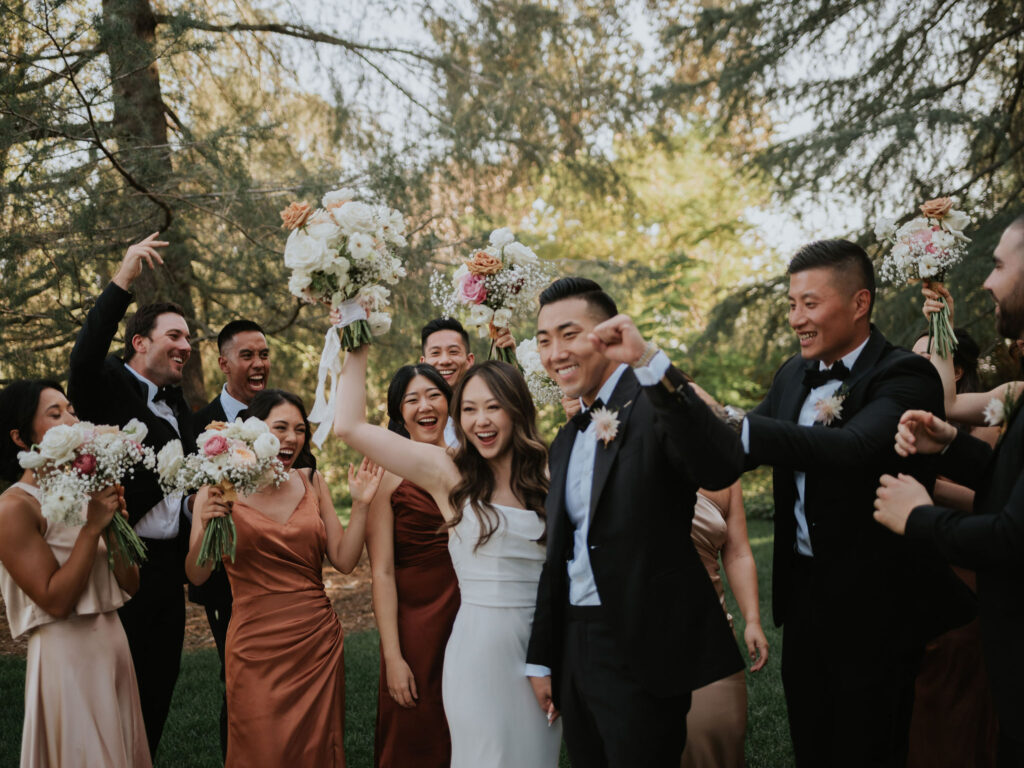 A joyful wedding party celebrating outdoors, with the bride and groom in the center, surrounded by bridesmaids and groomsmen holding bouquets.