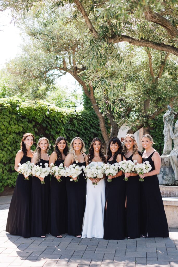 Bride and seven bridesmaids in black dresses pose with bouquets in a garden setting.