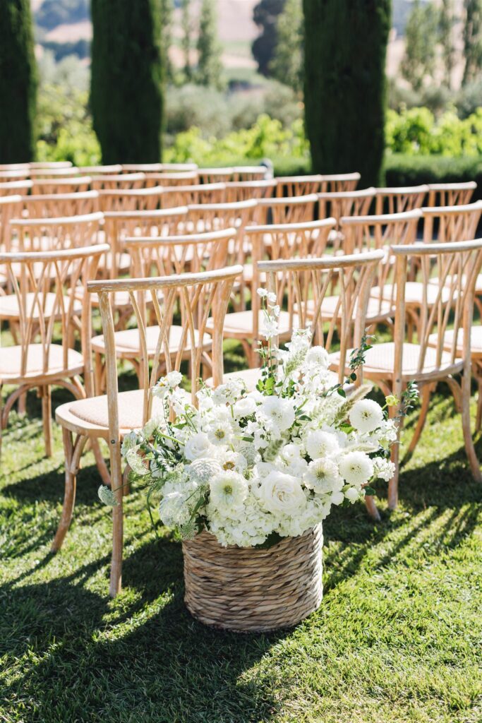Rows of wooden chairs are set up outdoors on grass, with a basket of white flowers placed at the end of an aisle.