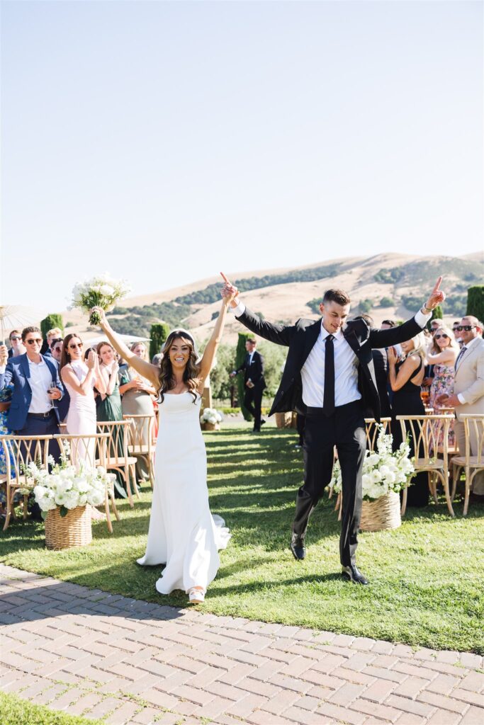A couple joyfully walks down the aisle outdoors, with guests clapping in the background. The bride holds a bouquet.