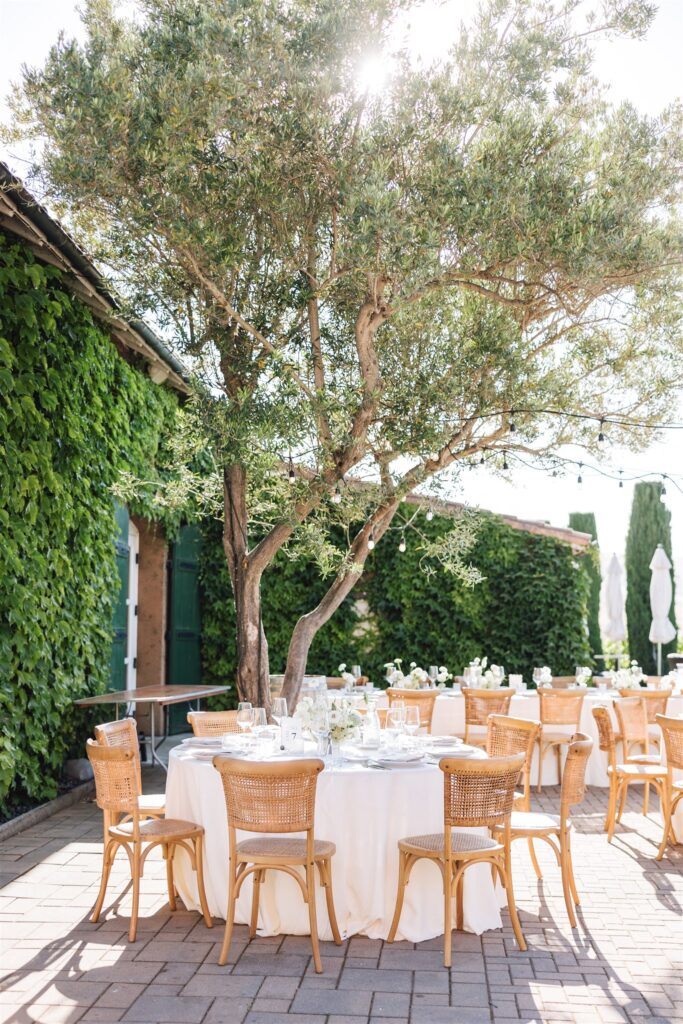 An outdoor dining setup with round tables and rattan chairs under a tree, surrounded by greenery-covered walls.