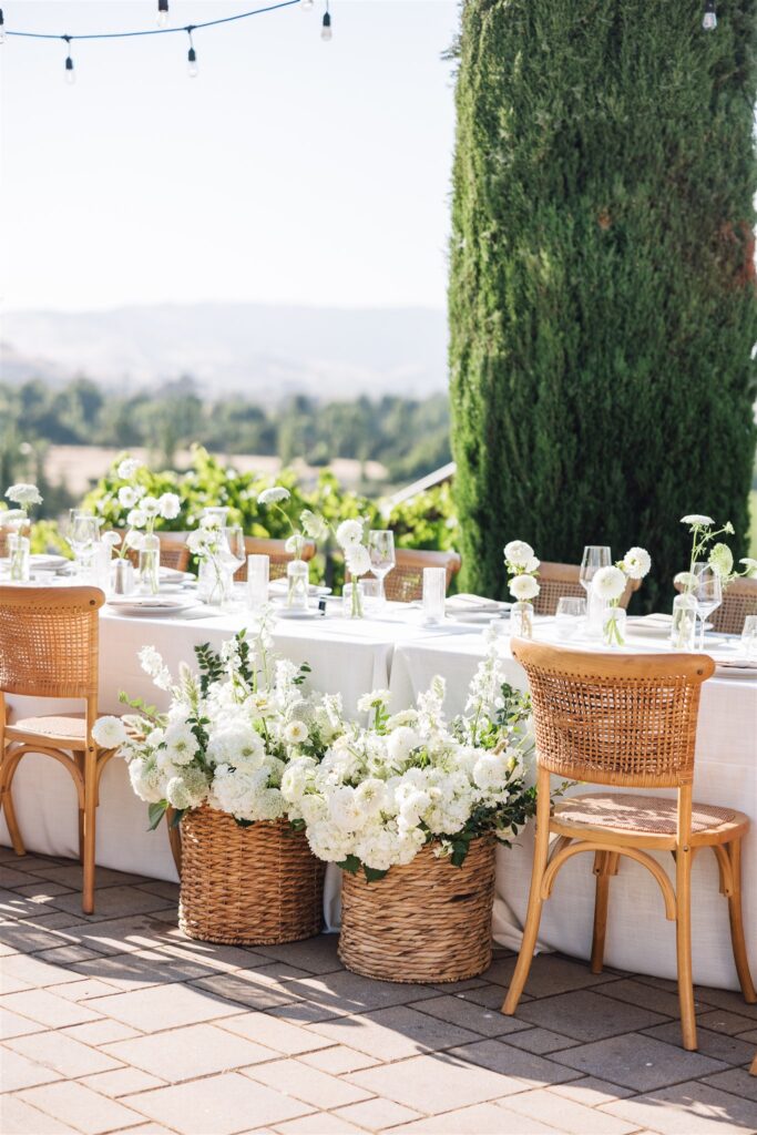 Outdoor dining setup with a long table, wicker baskets of white flowers, wooden chairs, and string lights. A tree and landscape are visible in the background.