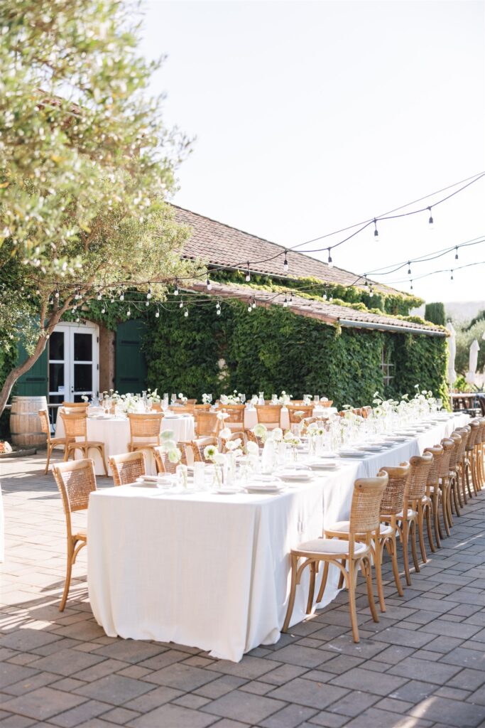 Outdoor dining setup with long tables covered in white tablecloths, wooden chairs, floral centerpieces, and string lights. Greenery and a vine-covered building are in the background.