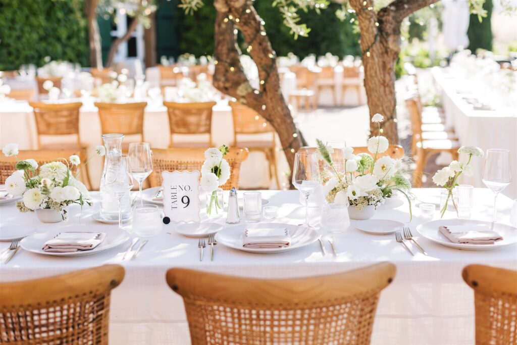 Elegant outdoor wedding reception with tables set for guests, featuring white flowers, glassware, and china, beneath string lights on trees.