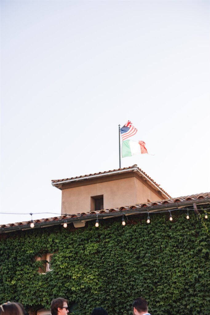Vine-covered building with string lights and two flags, American and Italian, on the roof against a clear sky.