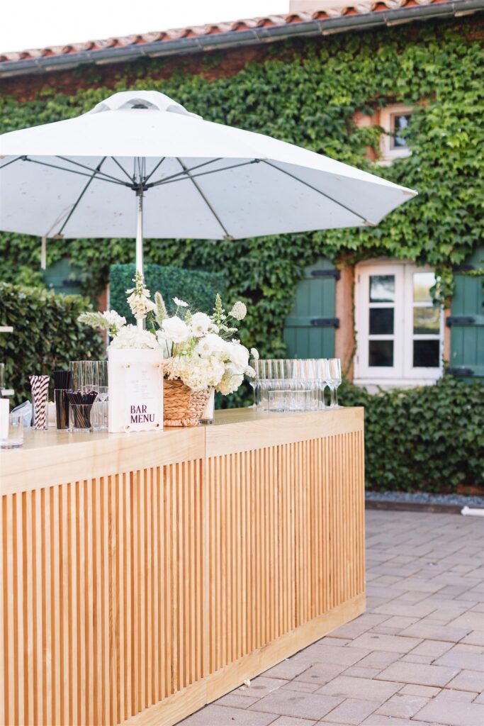 Outdoor bar setup with a wooden counter, white flowers, glassware, and a white umbrella, set against a building with green ivy and two shuttered windows.