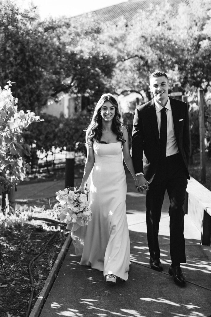 A couple in wedding attire holds hands and smiles while walking outdoors along a path, surrounded by greenery. Black and white photo.
