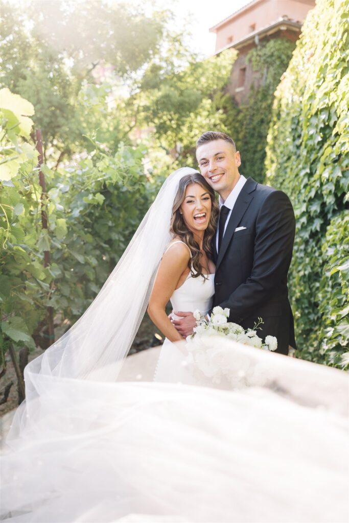 Bride and groom smiling in a garden setting, with the bride wearing a long veil and holding a bouquet.