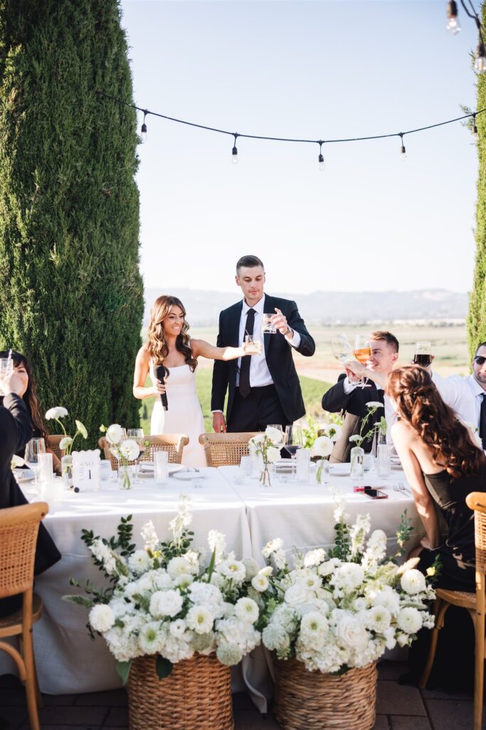 A wedding couple toasts with guests outdoors at a decorated table with white flowers, under string lights, against a scenic landscape backdrop.