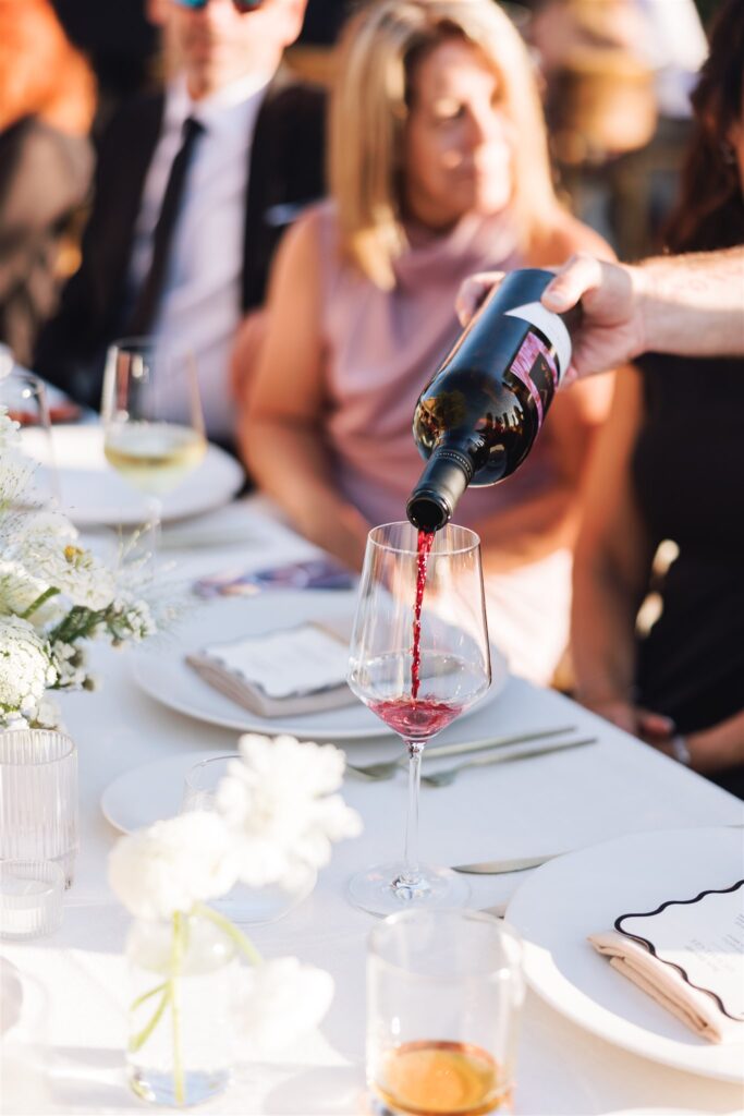 A person pours red wine into a glass on a table set for a meal, with plates, flowers, and menus visible.