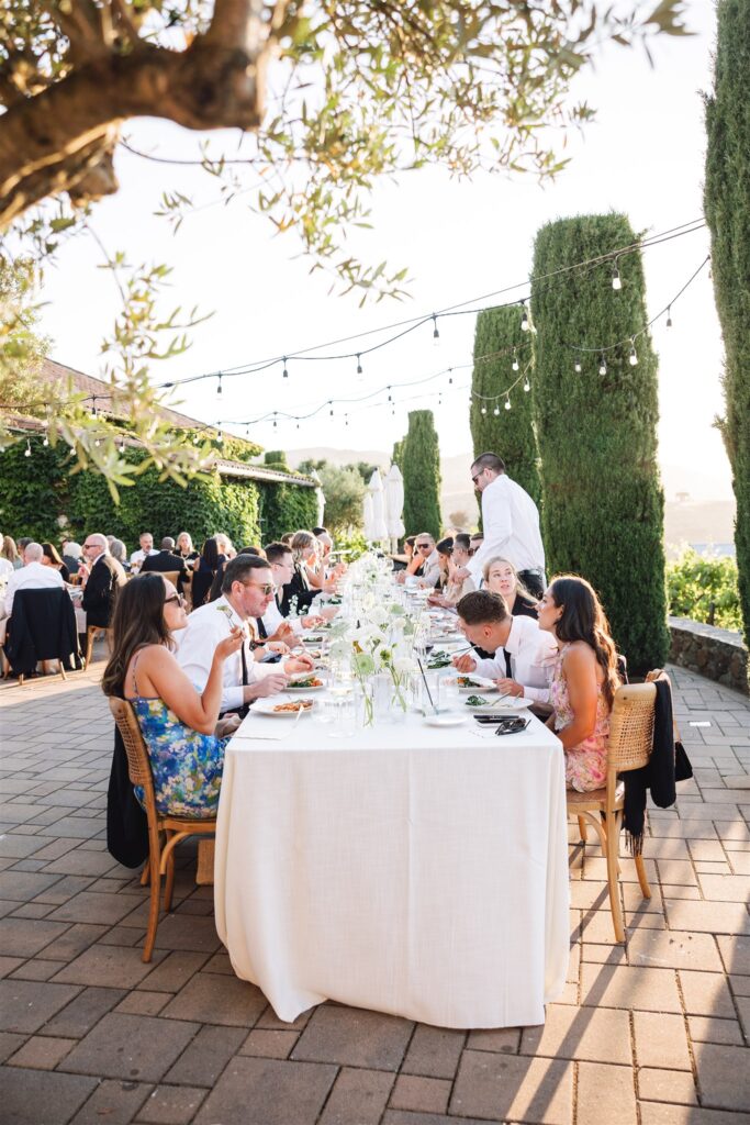 People seated around a long outdoor table, dining under string lights and surrounded by tall trees and greenery, with distant hills visible in the background.