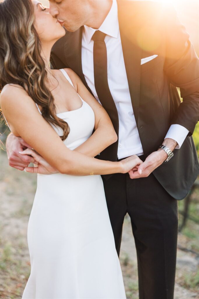 A couple in formal attire shares a kiss, standing closely with the sun shining brightly in the background.
