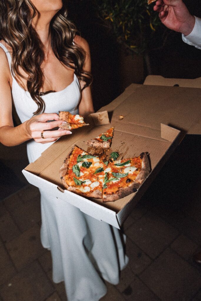 Person in white dress holding a pizza box with a sliced pizza, topped with basil and cheese, while another hand reaches for a slice.