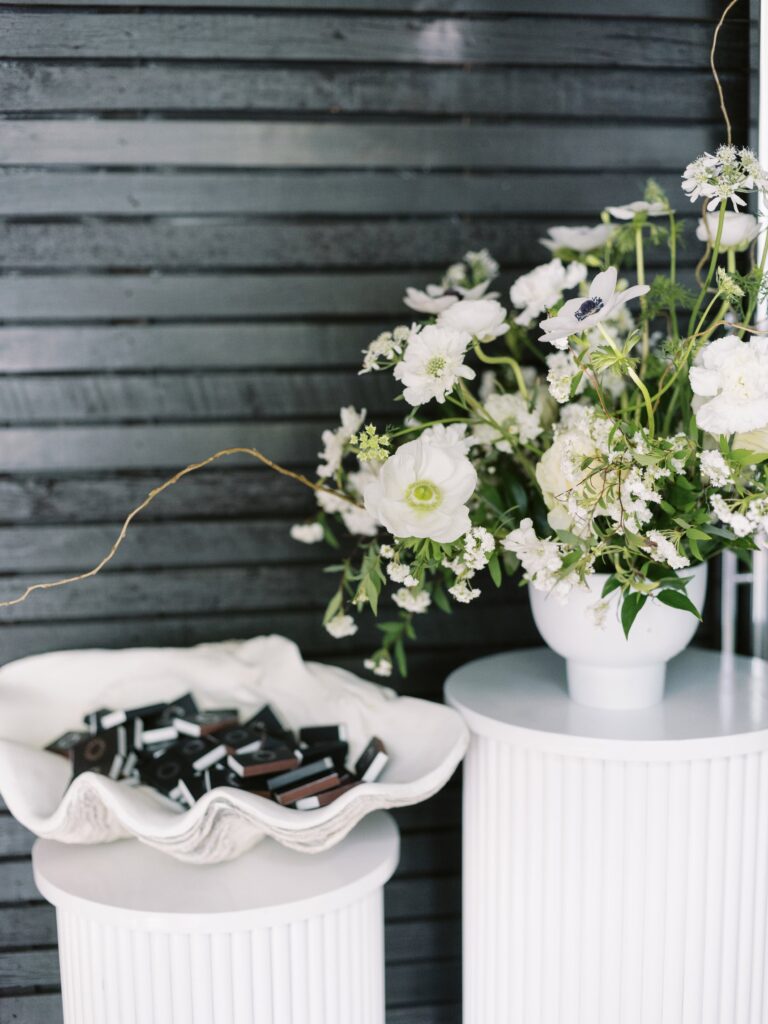 White floral arrangement in a vase on a round pedestal next to a shell-shaped dish holding small rectangular black objects. Background features a dark horizontal wooden slat wall.