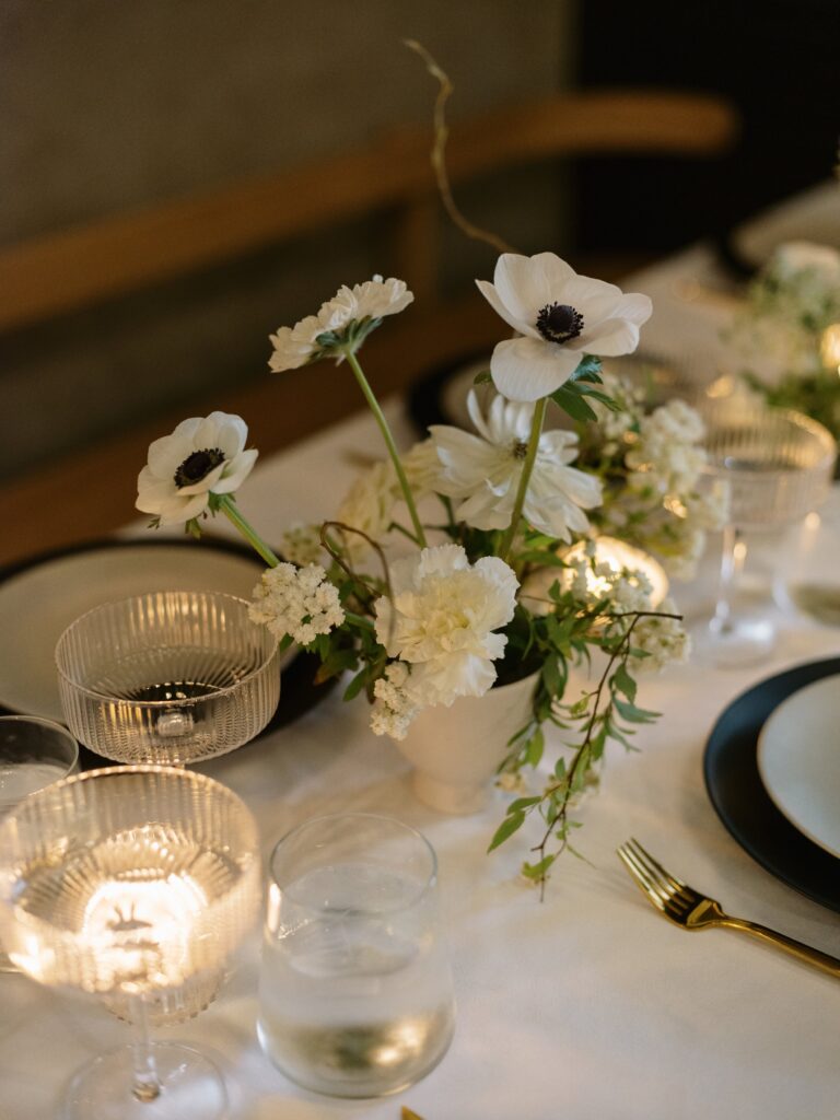 Elegant table setting with white flowers in a vase, surrounded by glassware and plates on a white tablecloth.