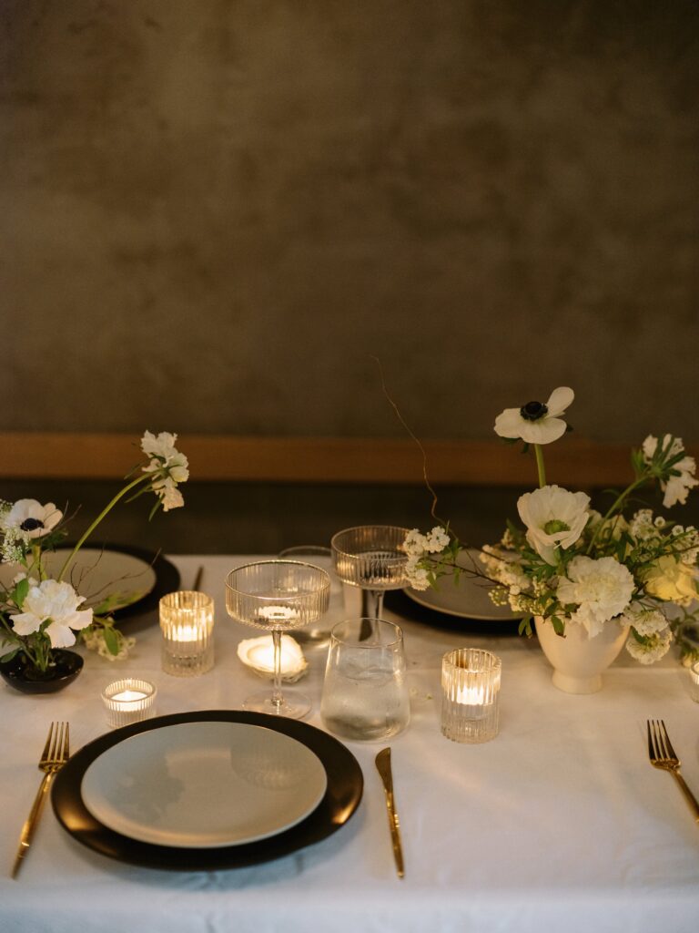 A dining table set with black-rimmed plates, glassware, lit candles, and white floral arrangements on a white tablecloth.