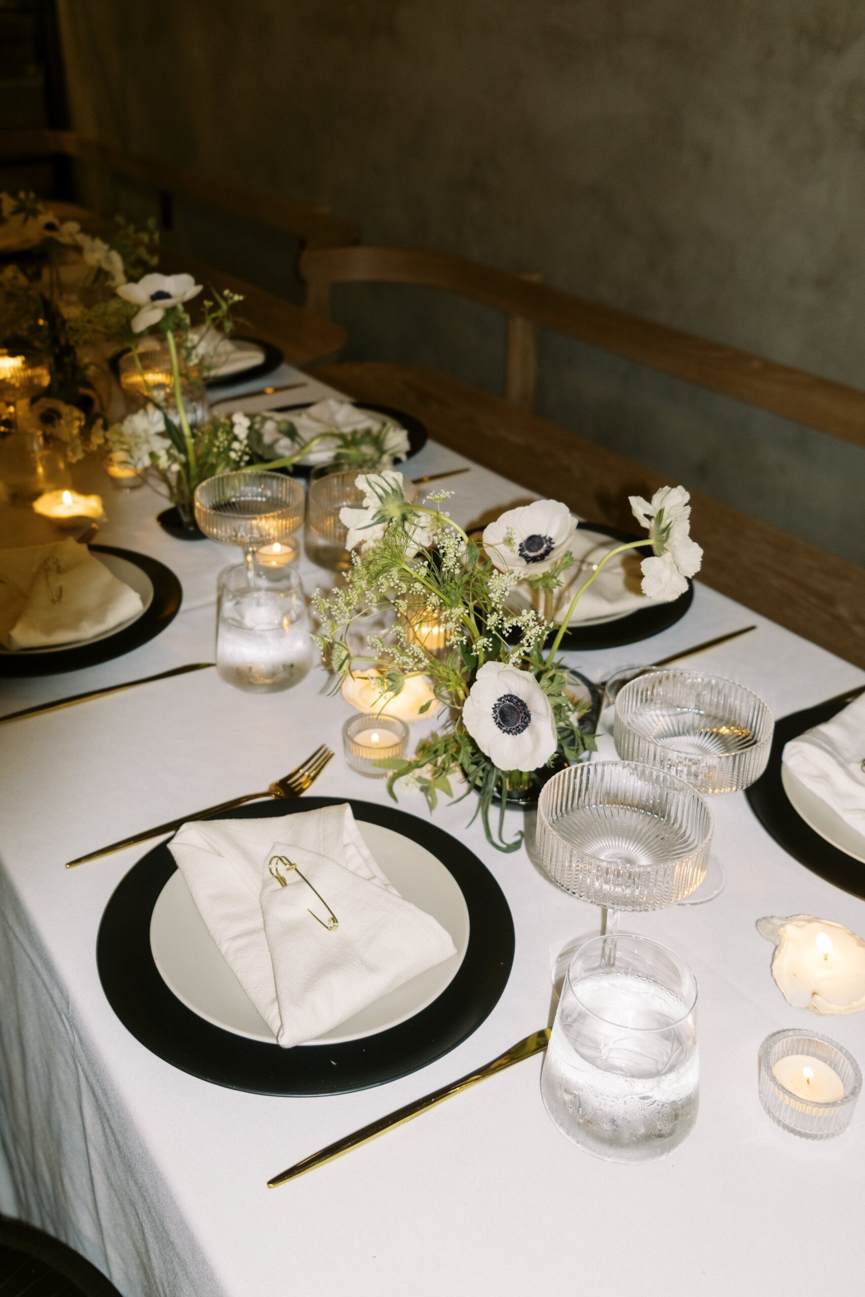 Elegant dining table set with black-rimmed plates, white napkins, glassware, candles, and floral centerpieces featuring white flowers.