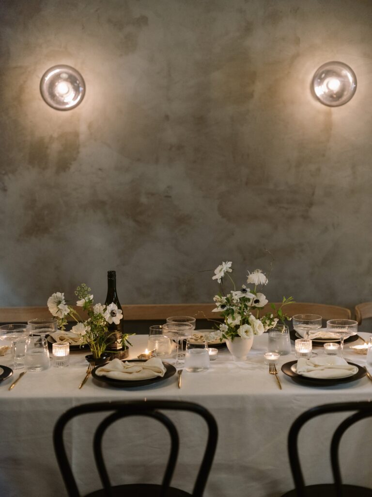 A dimly lit dining table with a white tablecloth, set with plates, glasses, candles, and floral arrangements. Two wall lights illuminate the scene.