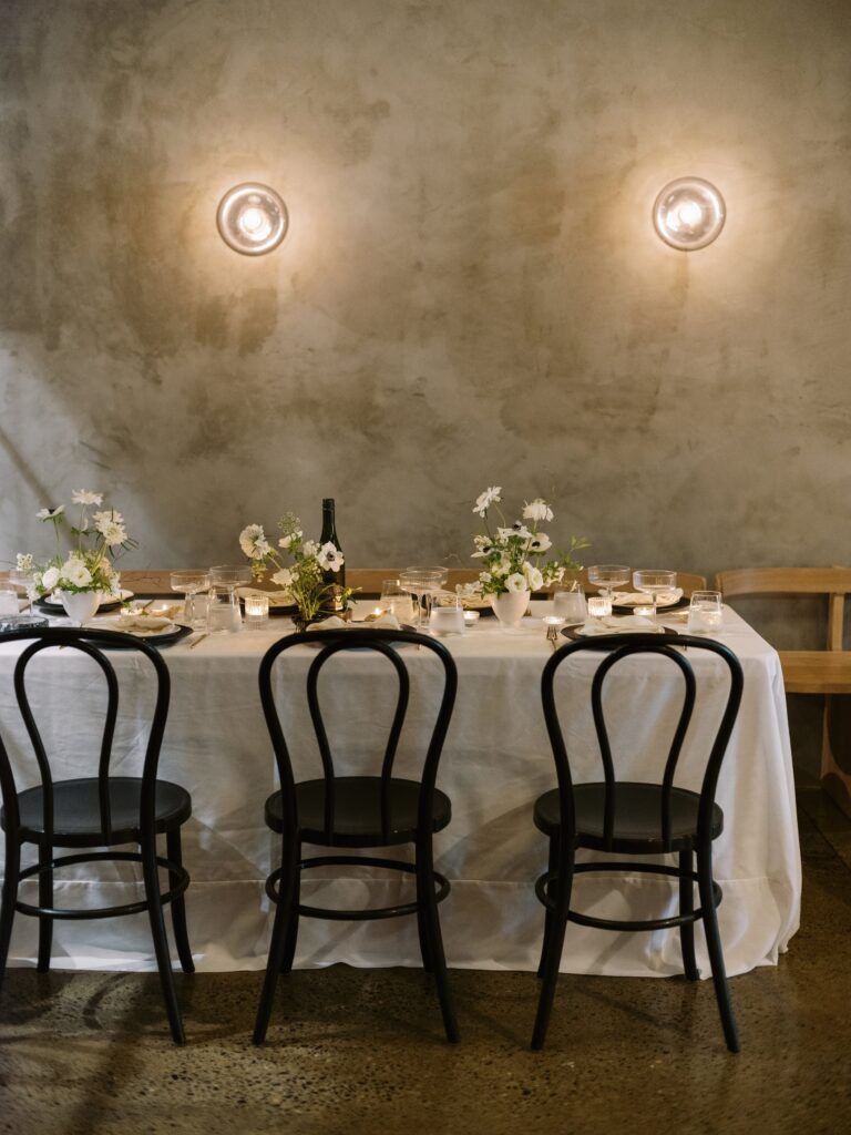 A dining table set for four with a white tablecloth, black chairs, flowers, glassware, and a bottle, against a textured gray wall with two round lights.