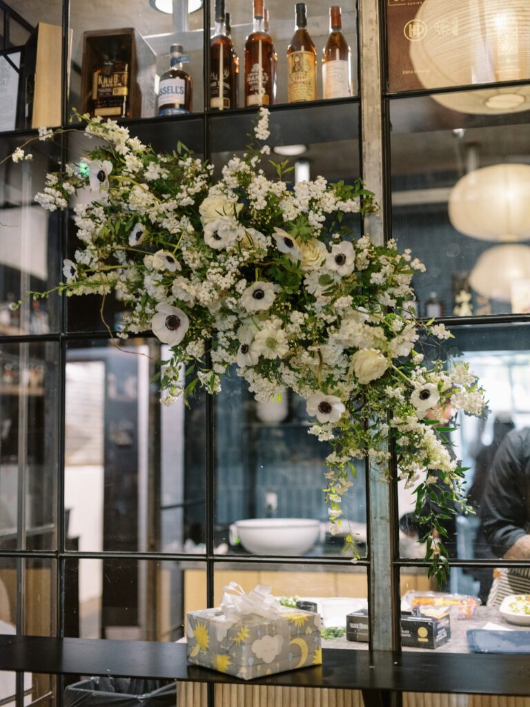 A floral arrangement with white flowers hangs on a grid wall above a table displaying a gift box. Bottles and decor elements are visible in the background.