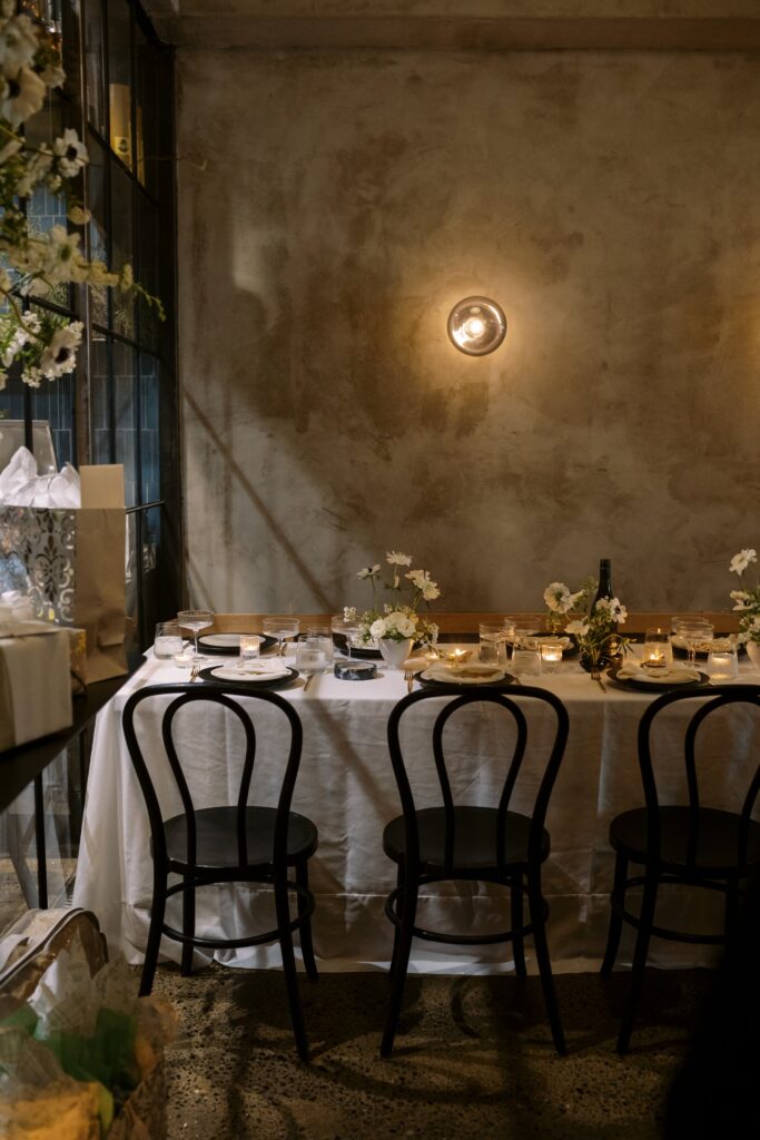 A dimly lit dining area with a table set for a meal, featuring white tablecloth, black chairs, candles, flowers, and a concrete wall in the background.